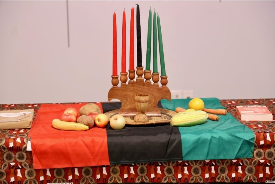 A kinara with red, black and green candles on a table set with fruit and a pan-African flag
